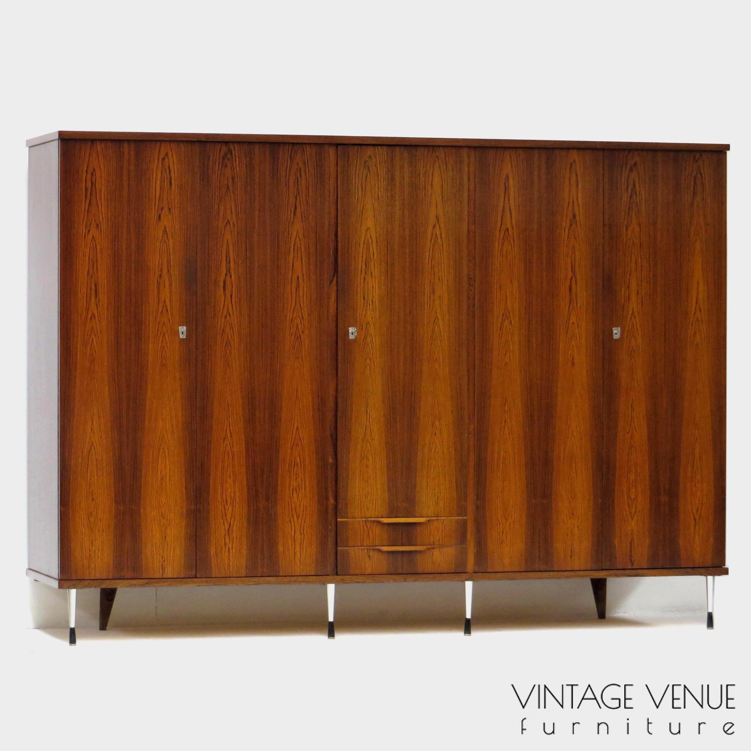 EN: 5-door vintage designer wardrobe in high quality rosewood rosewood, with stylish adjustable legs, 2 drawers with pretty handles, 4 shelves, 2 brass hanging rails, a mirror, and very sturdy adjustable & stylish metal legs. Period: 1960s. Condition: fully functional working and generally in good condition. Slight signs of use commensurate with age. ENG: Rare and exclusive high quality vintage mid century modern design wardrobe made of rosewood / rosewood. It has 5 doors, 2 drawers with beautiful carved handles, 4 shelves, 2 brass hang supports, a mirror and adjustable stylish and very solid metal legs. Period: 1960s. Condition: fully functional and overall in good condition. Light wear consistent with age and use. EN: 5-door vintage designer wardrobe in high quality rosewood rosewood, with stylish adjustable legs, 2 drawers with pretty handles, 4 shelves, 2 brass hanging rails, a mirror, and very sturdy adjustable & stylish metal legs. Period: 1960s. Condition: fully functional working and generally in good condition. Slight signs of use commensurate with age. ENG: Rare and exclusive high quality vintage mid century modern design wardrobe made of rosewood / rosewood. It has 5 doors, 2 drawers with beautiful carved handles, 4 shelves, 2 brass hang supports, a mirror and adjustable stylish and very solid metal legs. Period: 1960s. Condition: fully functional and overall in good condition. Light wear consistent with age and use.